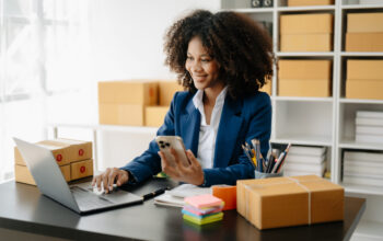 Startup small business SME, Entrepreneur owner African woman usi Startup small business SME, Entrepreneur owner African woman using smartphone or tablet taking receive and checking online purchase shopping order to preparing pack product box. in office