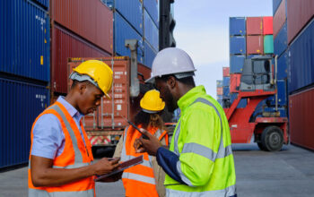 Black male workers and teams work at a logistic crane with stacks of containers. African American male workers and teams in safety uniforms and hardhats use walkie-talkies, work at logistic crane with stacks of containers, load control shipping goods, and cargo transport industry.