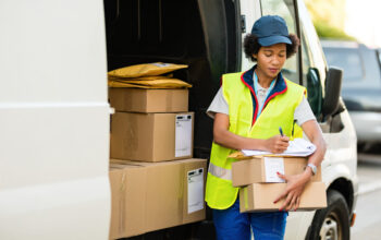 African American courier writing while checking deliveries by he Black delivery woman filling shipment list while standing with packages next to a mini van.