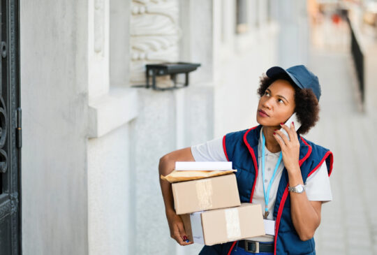 African American female deliverer carrying packages and talking on mobile phone in the city.