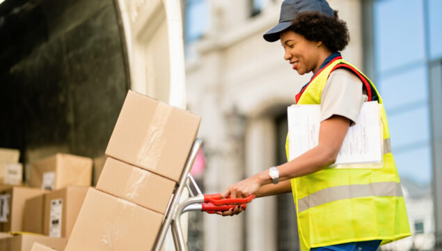 Happy black courier using hands truck while loading packages in a delivery van.
