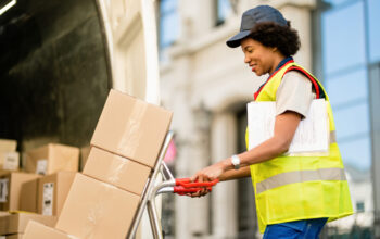 Happy black courier using hands truck while loading packages in a delivery van.