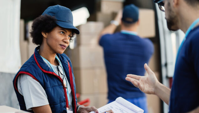 Two deliverers communicating and going through paperwork while their colleagues is loading packages in a van. Focus is on African American woman.
